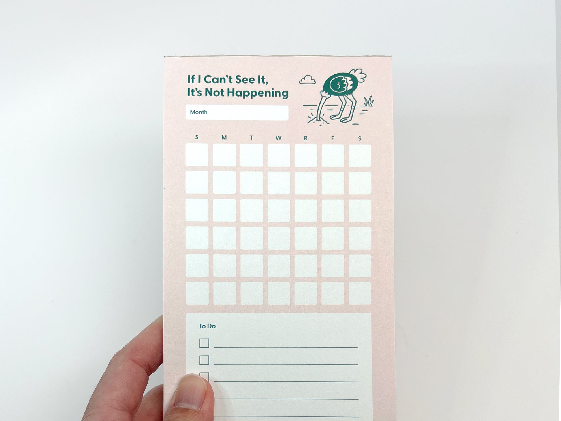 Front-facing photo of a tall, slim monthly planner notepad held near the bottom edge against a white background. The notepad has a soft pink background with the phrase “If I Can’t See It, It’s Not Happening” at the top and a small illustrated ostrich with its head in the ground. Below the title is a blank month label, a full monthly calendar grid with weekday initials, and a lined to-do list section partially visible at the bottom.