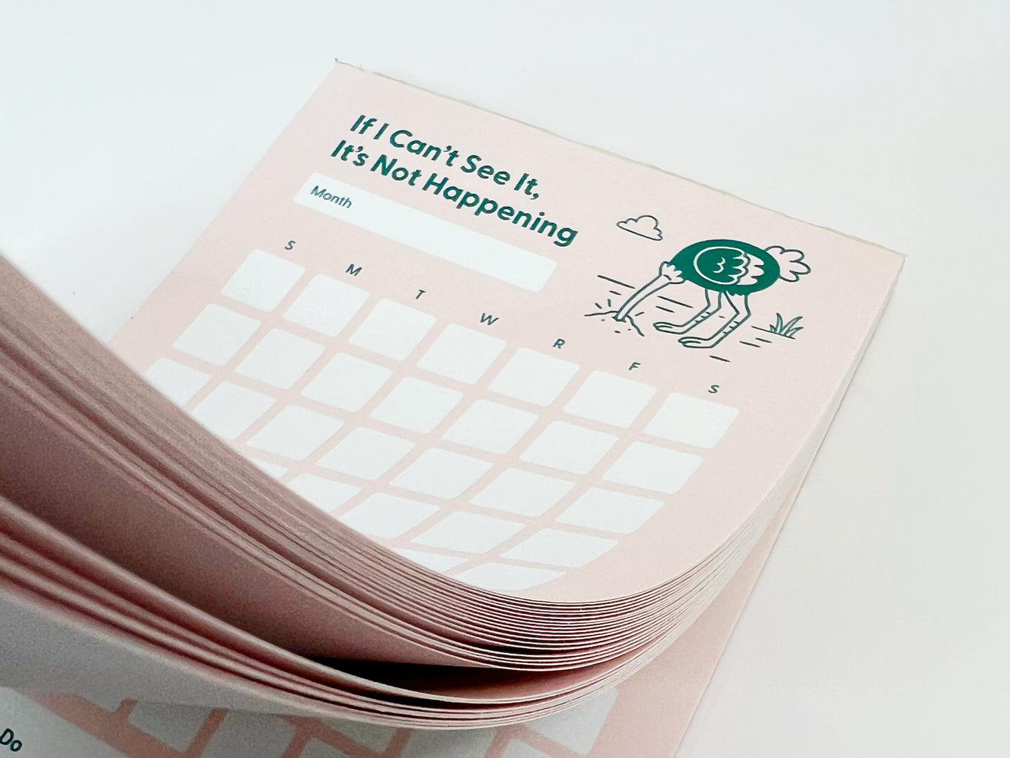 Close-up photo of a thick stack of a tall monthly planner notepad with soft pink pages slightly fanned. The top sheet reads “If I Can’t See It, It’s Not Happening” and shows a blank month label, a monthly calendar grid with weekday initials, and a small illustrated ostrich with its head in the ground. The layered paper edges are clearly visible against a white background.