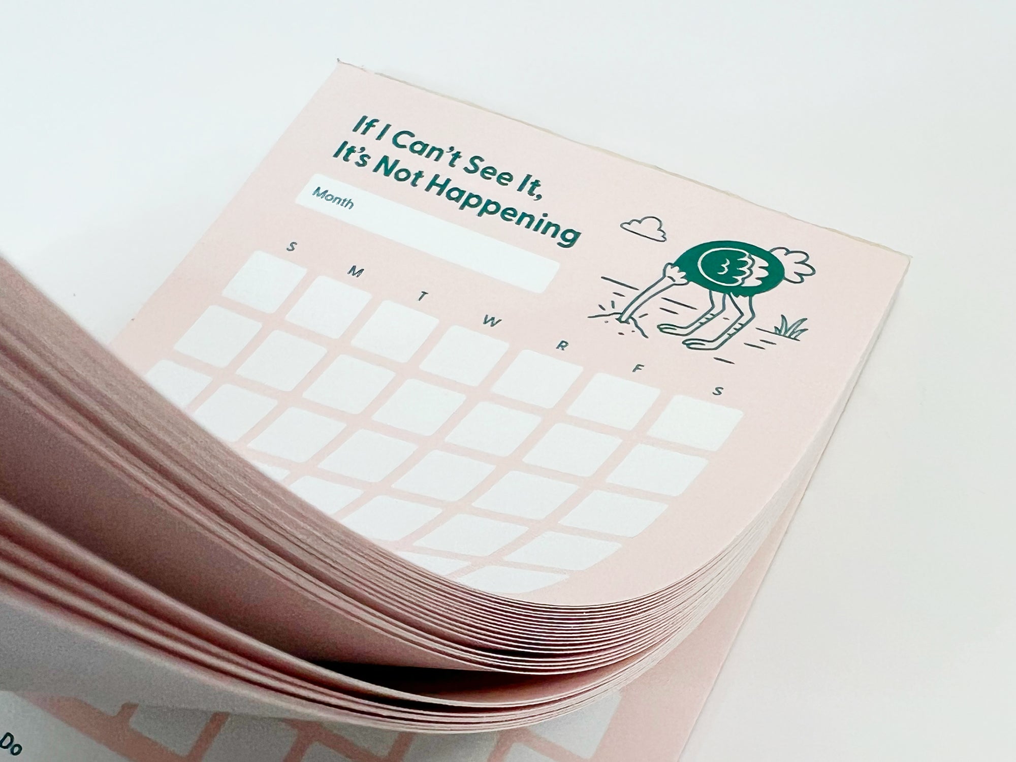 Close-up photo of a thick stack of a tall monthly planner notepad with soft pink pages slightly fanned. The top sheet reads “If I Can’t See It, It’s Not Happening” and shows a blank month label, a monthly calendar grid with weekday initials, and a small illustrated ostrich with its head in the ground. The layered paper edges are clearly visible against a white background.