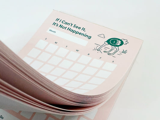 Close-up photo of a thick stack of a tall monthly planner notepad with soft pink pages slightly fanned. The top sheet reads “If I Can’t See It, It’s Not Happening” and shows a blank month label, a monthly calendar grid with weekday initials, and a small illustrated ostrich with its head in the ground. The layered paper edges are clearly visible against a white background.