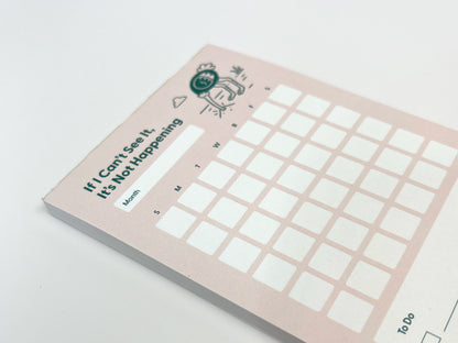 Angled overhead photo of a tall monthly planner notepad lying flat on a white surface. The notepad has a soft pink background with the phrase “If I Can’t See It, It’s Not Happening” at the top and a small illustrated ostrich with its head in the ground. A blank month label and a full monthly calendar grid with weekday initials are visible, with the thick paper stack shown along the bottom edge.