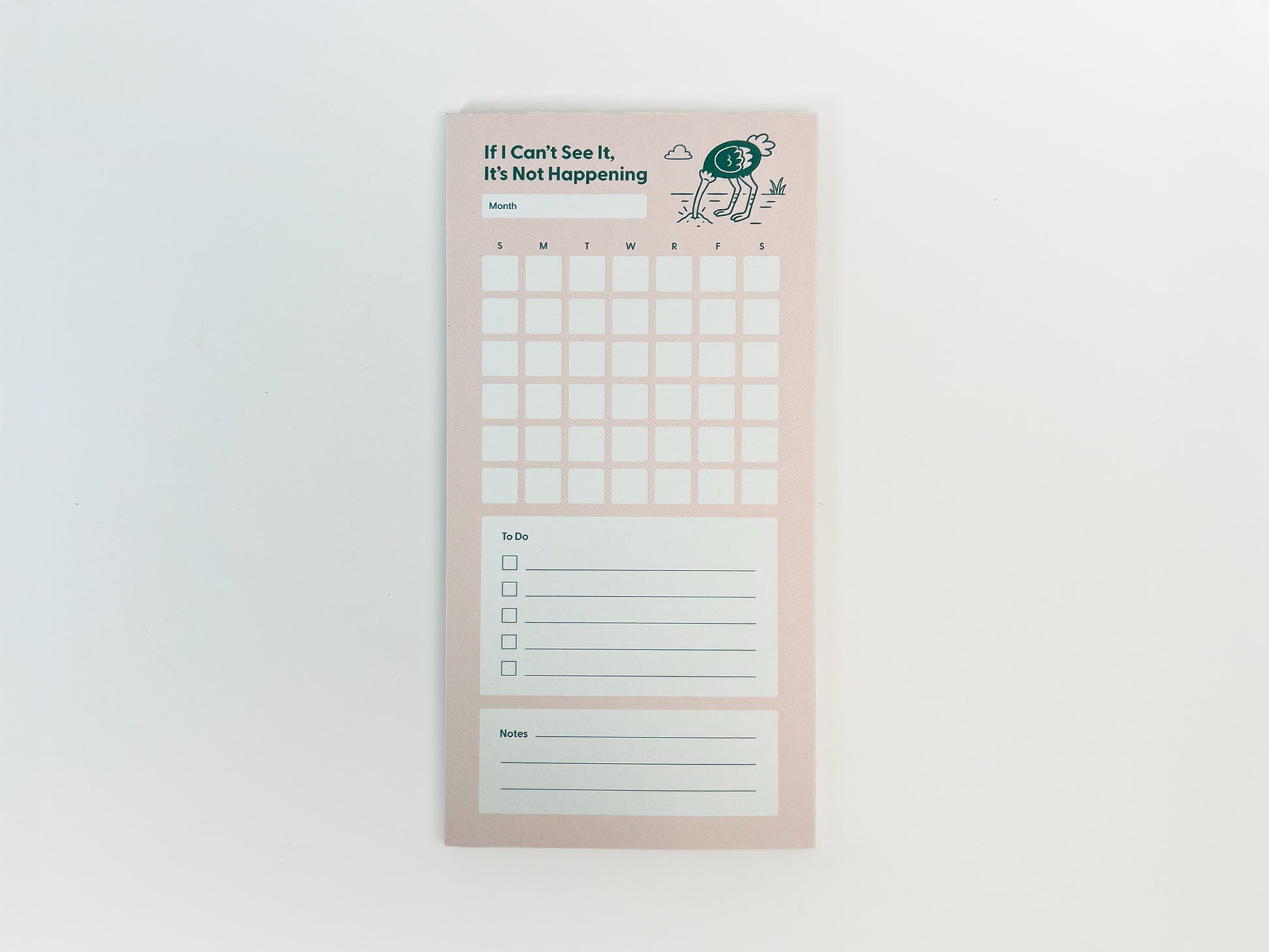 Full-length front-facing photo of a tall, narrow monthly planner notepad centered on a white background. The notepad has a soft pink background with the phrase “If I Can’t See It, It’s Not Happening” at the top and a small illustrated ostrich with its head in the ground. It features a blank month label, a full monthly calendar grid with weekday initials, a lined to-do list with checkboxes, and a lined notes section at the bottom.