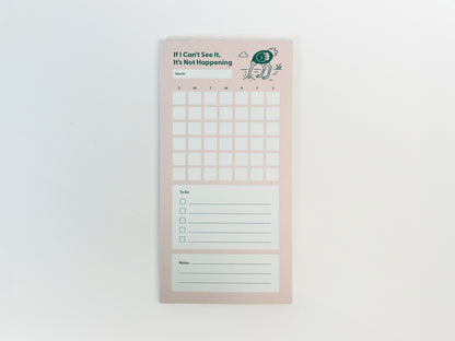 Full-length front-facing photo of a tall, narrow monthly planner notepad centered on a white background. The notepad has a soft pink background with the phrase “If I Can’t See It, It’s Not Happening” at the top and a small illustrated ostrich with its head in the ground. It features a blank month label, a full monthly calendar grid with weekday initials, a lined to-do list with checkboxes, and a lined notes section at the bottom.