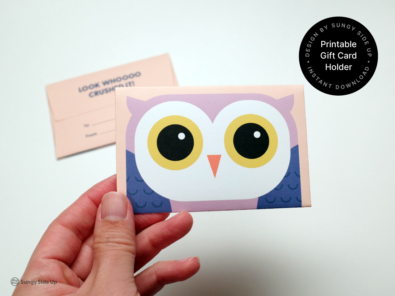 Hand holding a fully assembled printable owl gift card holder featuring a bold owl illustration with yellow eyes on a purple and peach envelope. A second holder with a “To and From” section is blurred in the background. This digital product is a fun and thoughtful option for gifting during graduations, school events, or thank you occasions. Easy-to-make and perfect for creative DIY gift presentations.