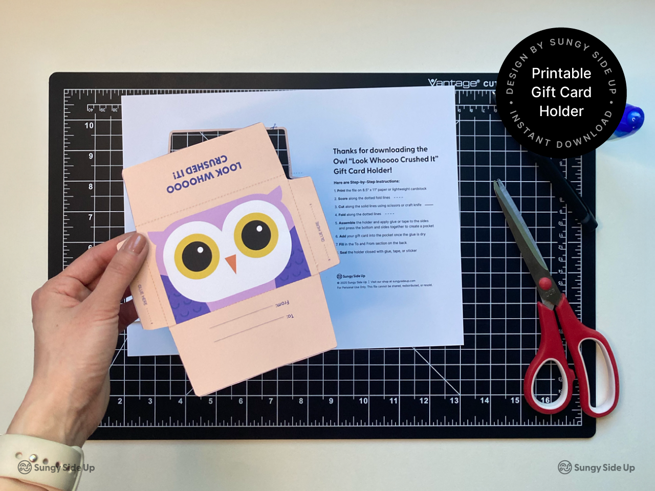 Hand holding a printable owl gift card holder above a printed instruction sheet on a black cutting mat. The peach and purple owl design is ready to be cut and assembled with scissors and glue. Perfect for a graduation gift or thank you note, this DIY instant download template is ideal for teachers, parents, and crafters seeking a fun printable envelope for standard-size gift cards.