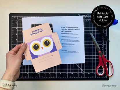 Hand holding a printable owl gift card holder above a printed instruction sheet on a black cutting mat. The peach and purple owl design is ready to be cut and assembled with scissors and glue. Perfect for a graduation gift or thank you note, this DIY instant download template is ideal for teachers, parents, and crafters seeking a fun printable envelope for standard-size gift cards.