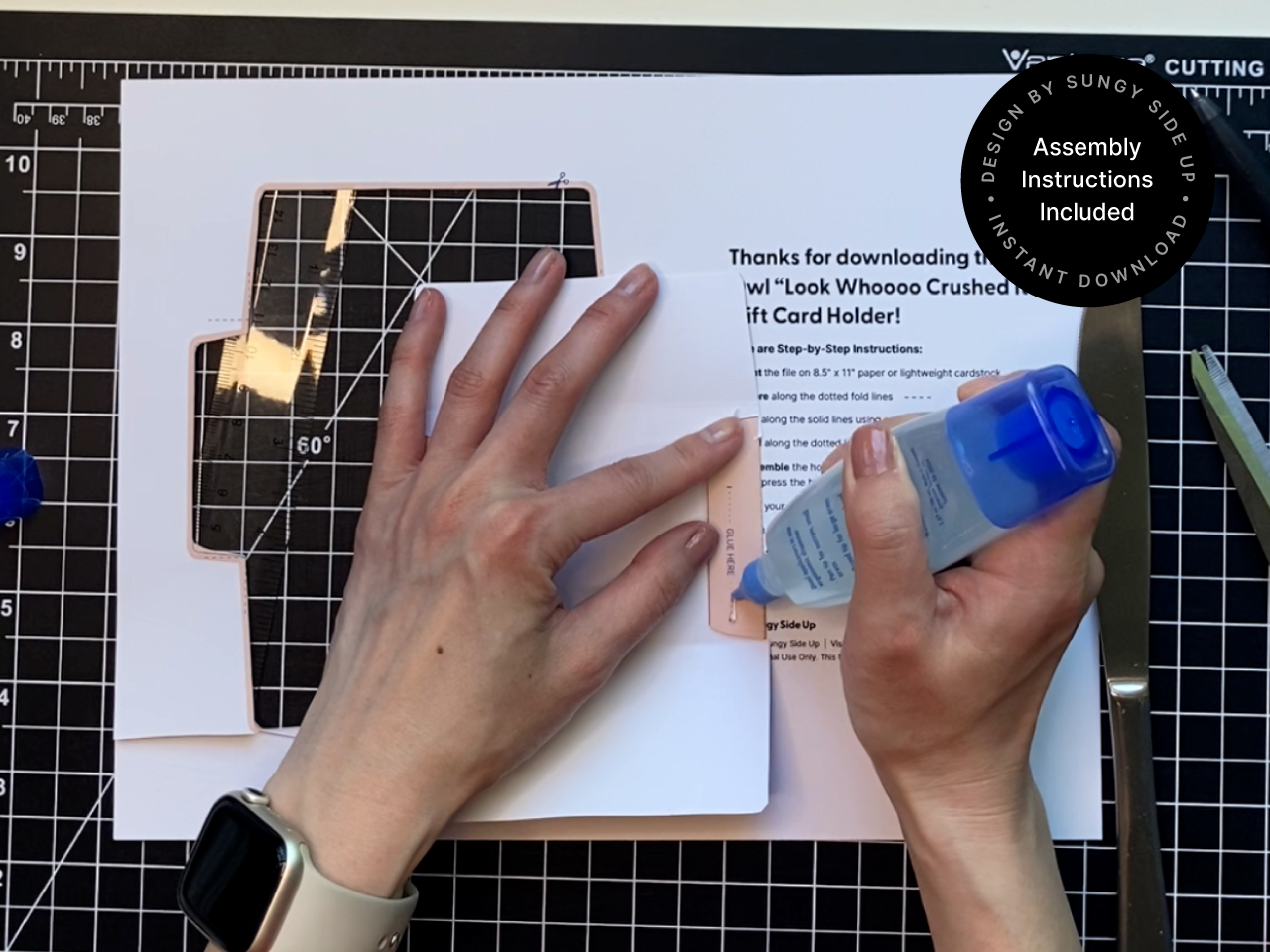 Hands applying liquid glue to the tab of a printable owl gift card holder template on a craft mat. The step-by-step printed instructions and scissors are visible in the background. This close-up shows how easy it is to assemble this digital download craft. Great for Shopify listings targeting DIY paper gifts, teacher appreciation, and graduation gift ideas.