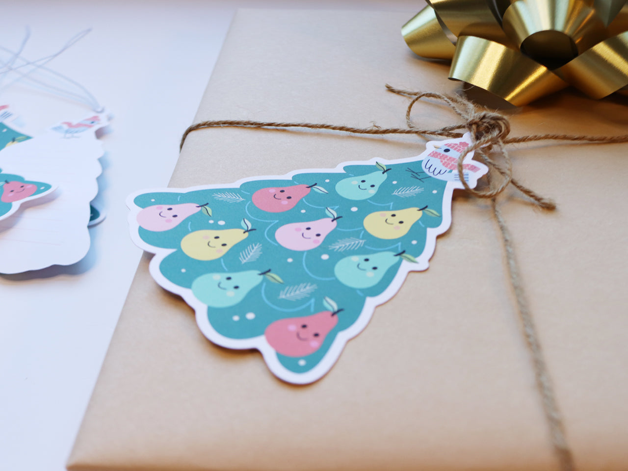 A close-up view of a kraft-wrapped gift tied with twine, showcasing a colorful Partridge in a Pear Tree tag with smiling pears and a bird illustration.