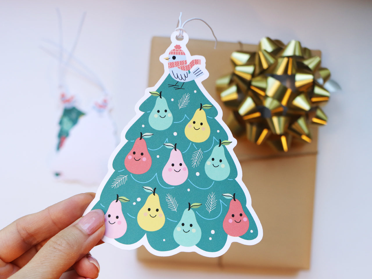 A hand holding a Christmas gift tag shaped like a tree, featuring a Partridge in a Pear Tree design with colorful smiling pears and a festive bird wearing a red hat and scarf. The background displays a kraft-wrapped gift with a golden bow.