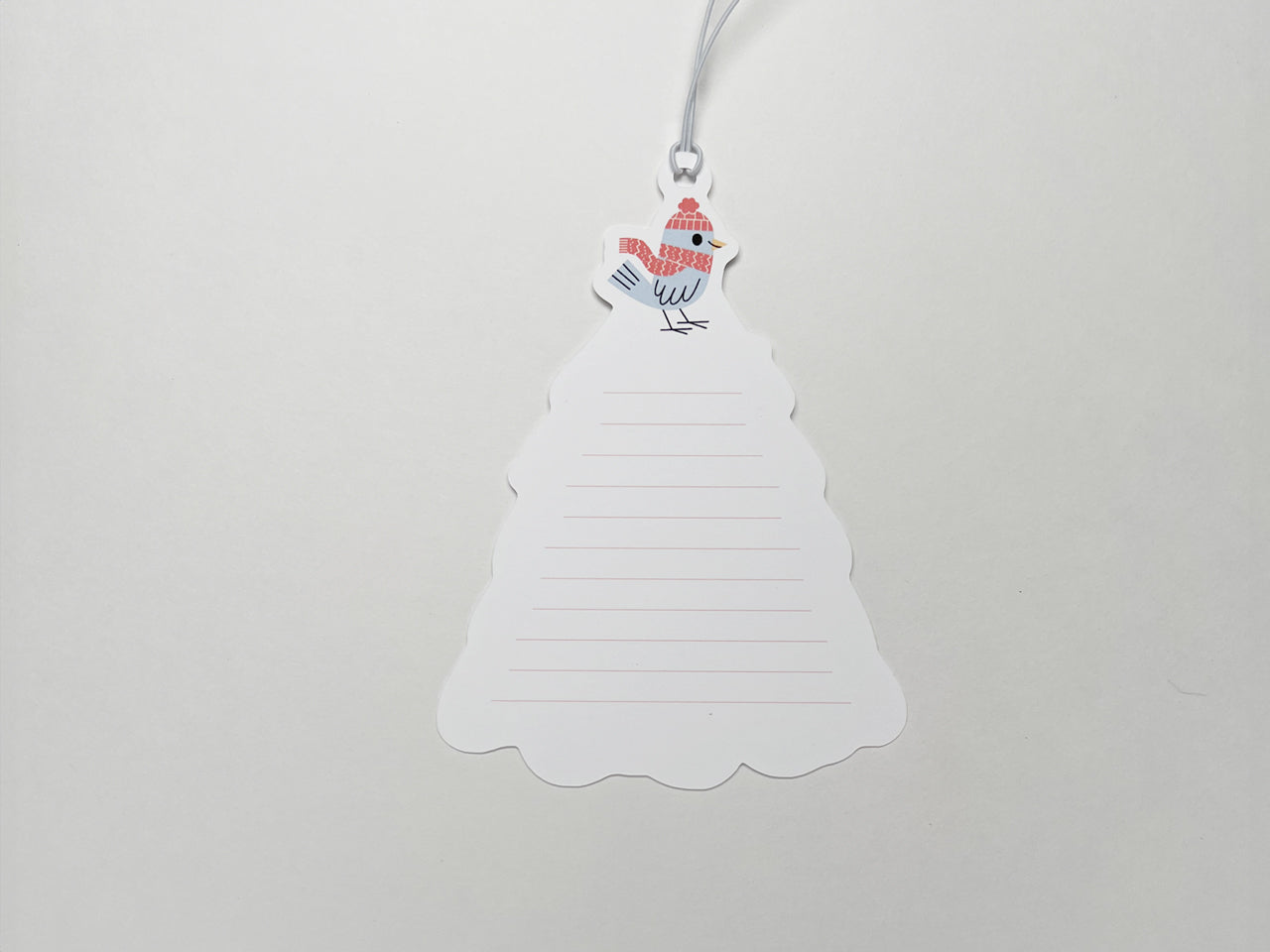 A close-up of the back of a Christmas gift tag shaped like a tree, with lined space for handwritten notes and a small bird illustration wearing a red winter hat and scarf at the top. Pre-attached white elastic string for easy attachment to gifts.