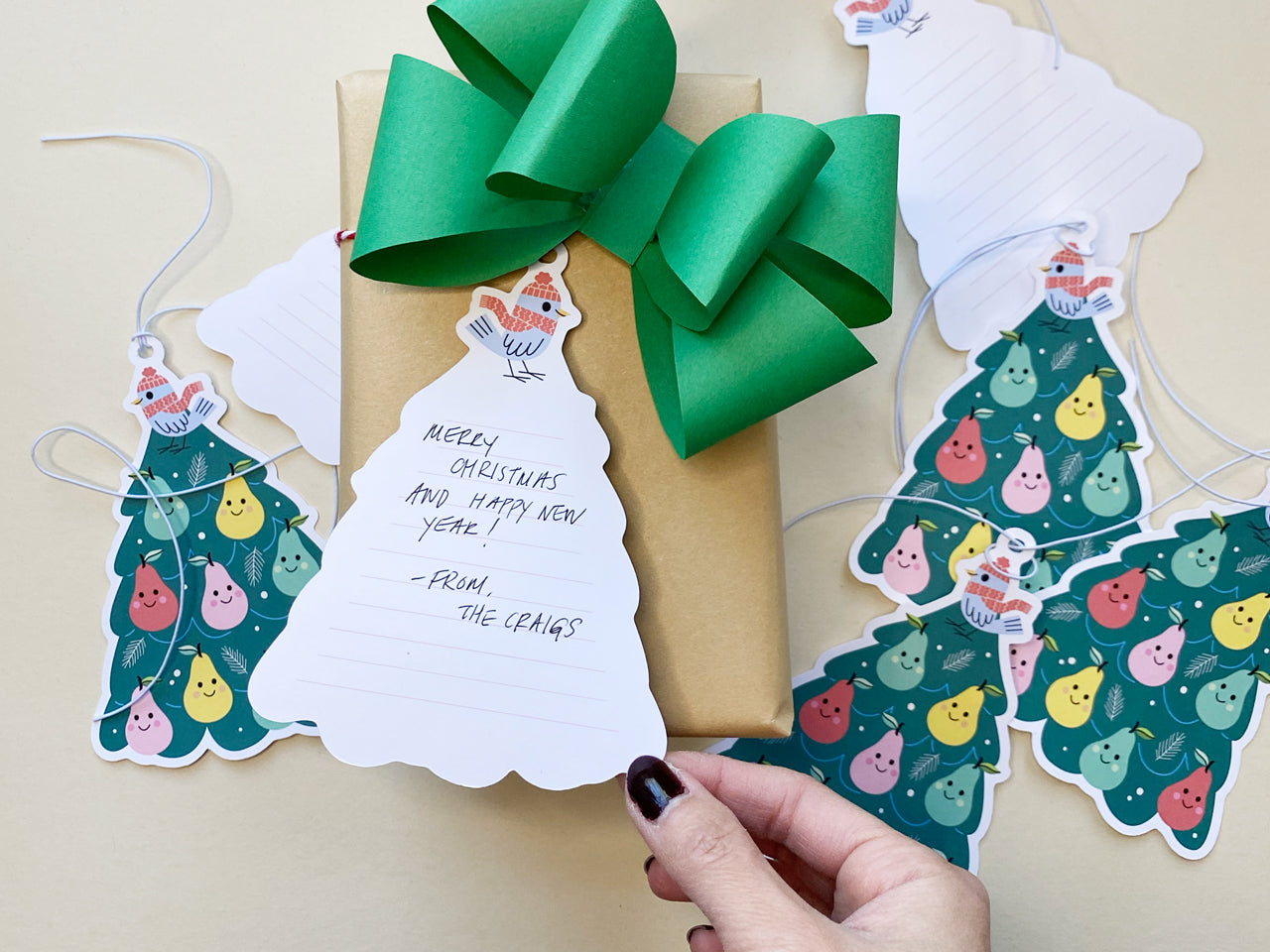 A person holding the back of a whimsical Partridge in a Pear Tree gift tag, handwritten with a holiday message on back. The die cut tag is shaped like a Christmas tree and features a bird wearing red hat and scarf illustration at the top.