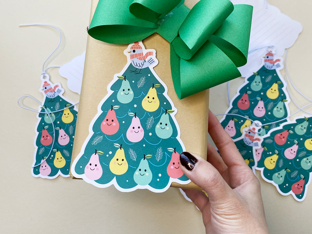 A hand holding a Christmas gift wrapped in kraft paper with a large green bow and a whimsical partridge in a pear tree gift tag. The gift tag features a colorful tree with smiling pears and a bird in a red hat and scarf.