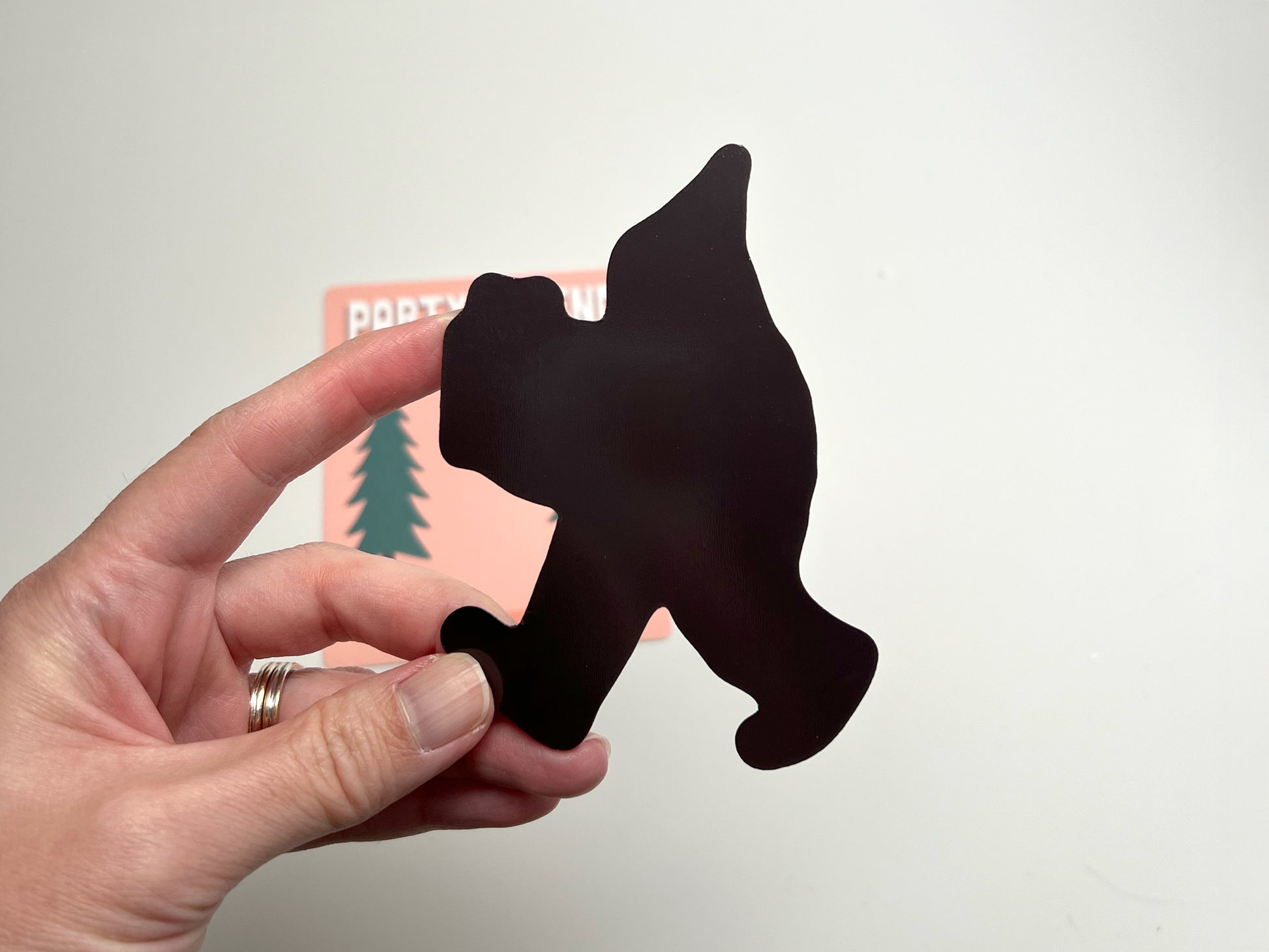 Back view of the Party Legend die-cut magnet shaped like a walking Sasquatch. The black magnetic side is shown being held up by a hand, highlighting its sturdy cut and smooth finish. The silhouette clearly displays the figure’s party hat and stride, emphasizing the quality of the magnet. In the background, part of the original backing card with pine tree artwork and peach color is visible, showing the playful packaging design.