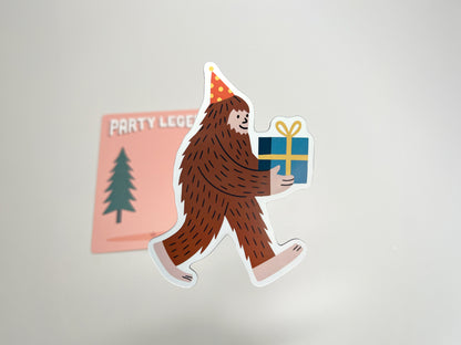 Close-up of the Party Legend die-cut magnet featuring a smiling Sasquatch wearing a red and yellow polka-dot party hat while carrying a wrapped blue gift with a yellow bow. The magnet is shown lifted slightly in front of its peach backing card, which has tree illustrations and “Party Legend” text. This quirky Bigfoot design makes a fun party favor, keepsake, or fridge decoration that adds playful, legendary charm to any celebration.