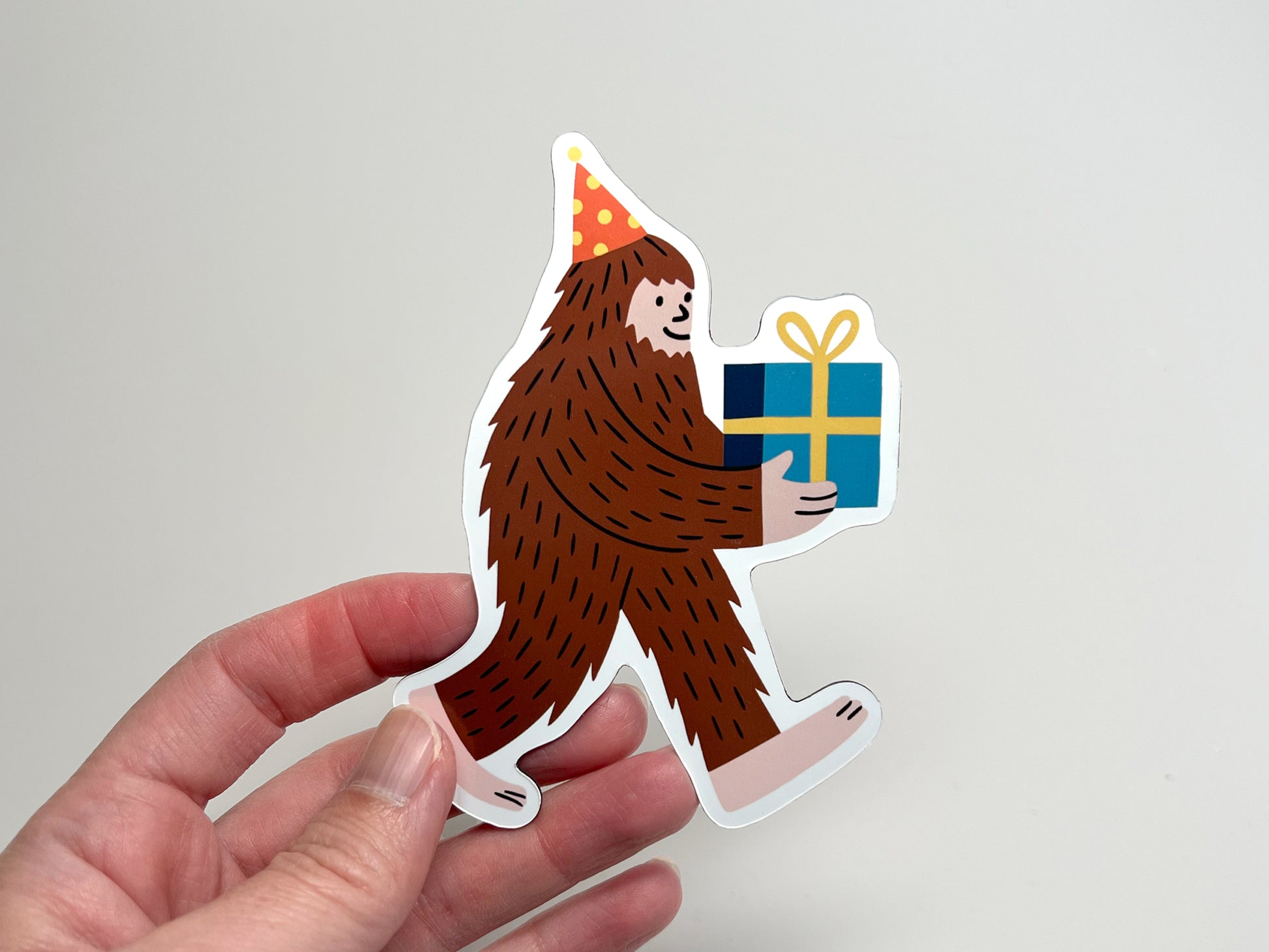Hand holding a die-cut Party Legend magnet featuring a cheerful Sasquatch wearing a polka-dot party hat and carrying a blue gift with a yellow bow. The quirky Bigfoot design makes a fun party favor, fridge decoration, or unique keepsake.