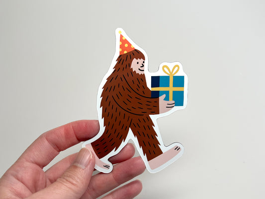 Hand holding a die-cut Party Legend magnet featuring a cheerful Sasquatch wearing a polka-dot party hat and carrying a blue gift with a yellow bow. The quirky Bigfoot design makes a fun party favor, fridge decoration, or unique keepsake.