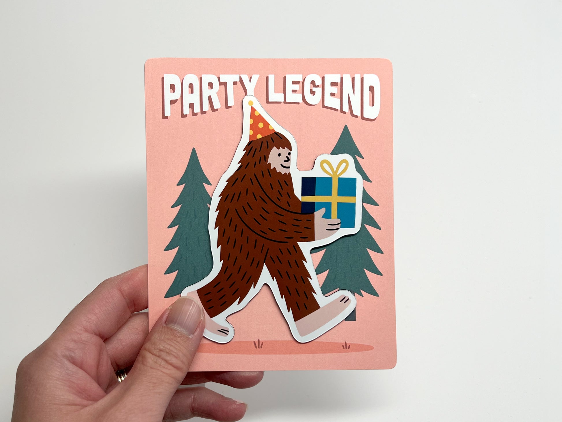 Die-cut Party Legend magnet on its backing card, featuring a fun Sasquatch design wearing a polka-dot party hat and carrying a wrapped gift with a yellow bow. The quirky Bigfoot illustration is set against pine trees with bold “Party Legend” text on a peach background. Ideal as a party favor, birthday keepsake, or funny fridge decoration, this handmade magnet adds playful charm and makes a unique gift for guests who love offbeat, legendary vibes.