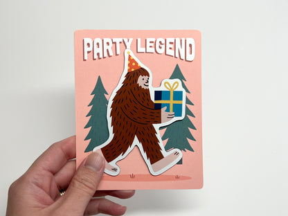 Die-cut Party Legend magnet on its backing card, featuring a fun Sasquatch design wearing a polka-dot party hat and carrying a wrapped gift with a yellow bow. The quirky Bigfoot illustration is set against pine trees with bold “Party Legend” text on a peach background. Ideal as a party favor, birthday keepsake, or funny fridge decoration, this handmade magnet adds playful charm and makes a unique gift for guests who love offbeat, legendary vibes.