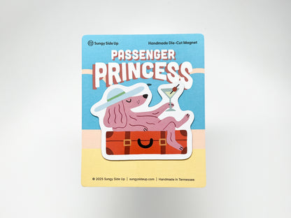Handmade die-cut magnet featuring a pink dog lounging on a suitcase with a martini, wearing a wide-brim hat. Backing card reads “Passenger Princess” in bold retro letters on a sunny travel-themed background. Fun, quirky gift for pet lovers, travelers, or anyone embracing the passenger princess vibe.