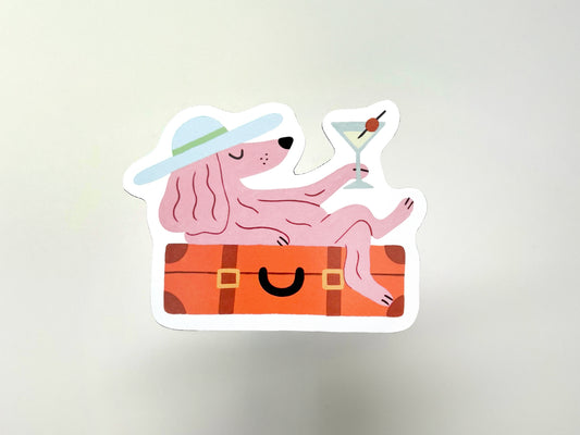 Handmade die-cut magnet featuring a pink dog lounging on an orange suitcase while holding a martini with an olive garnish. The dog wears a wide-brim hat with a green band, giving off relaxed vacation vibes. Fun, quirky design perfect for pet lovers, travelers, or anyone embracing their “passenger princess” era.