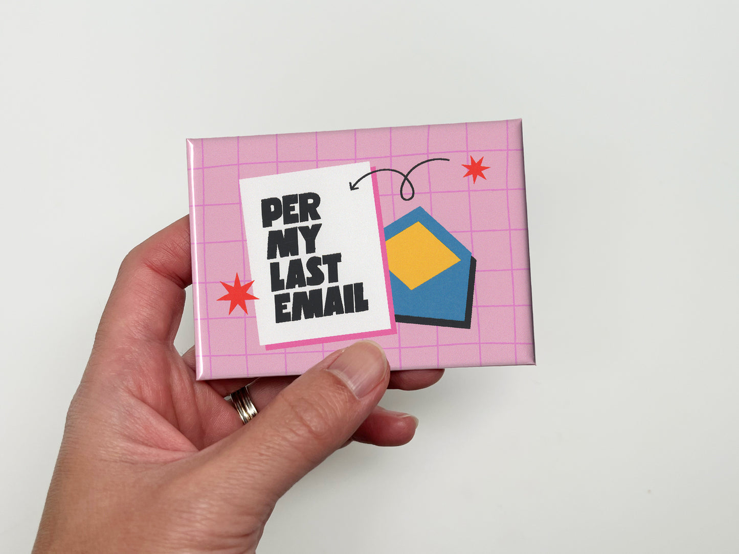 Hand holding a glossy pink magnet with a light pink grid background, bold black text that reads “Per My Last Email,” and a blue envelope illustration with red star accents.
