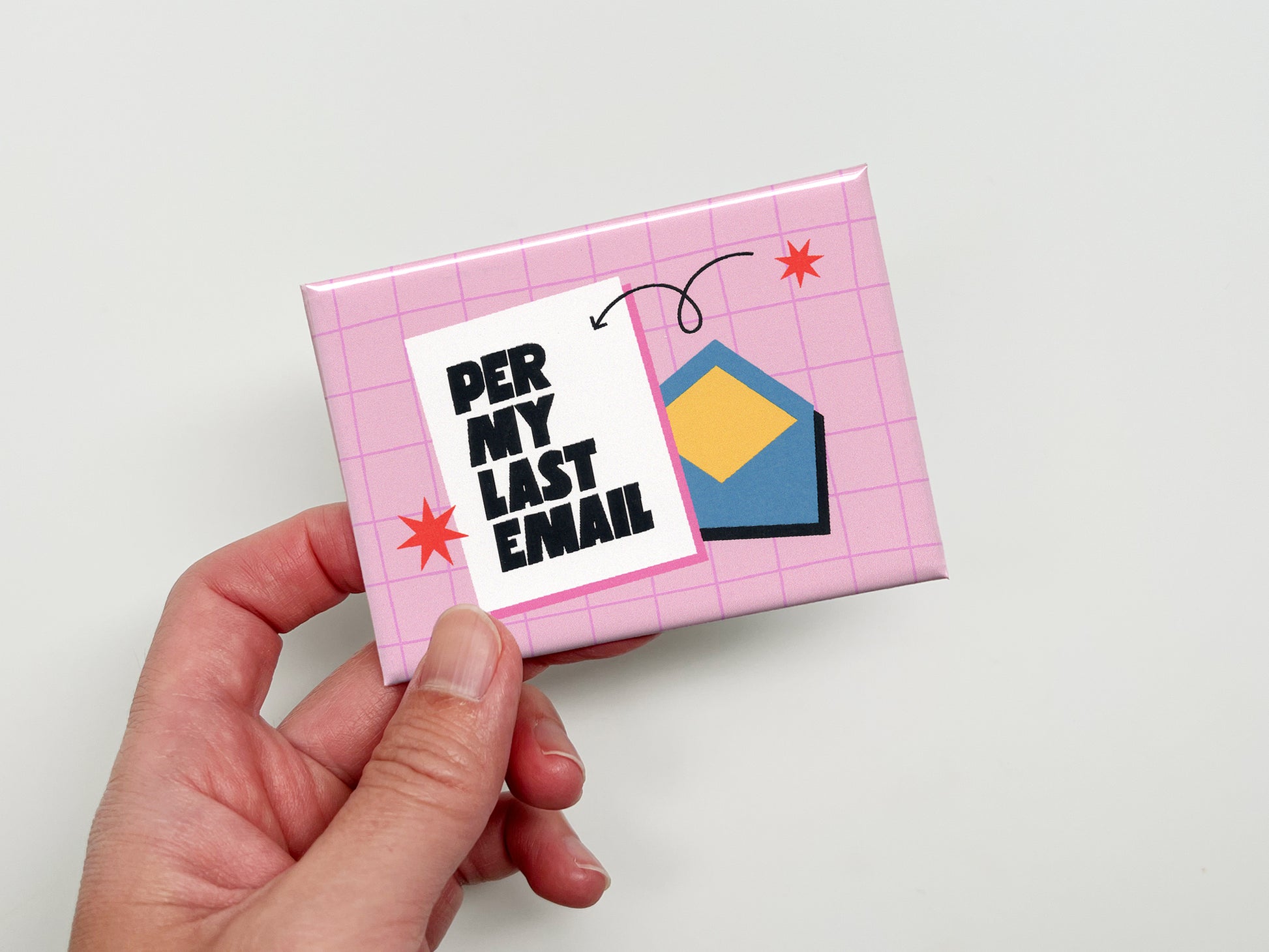 Hand holding a rectangular pink magnet with a light pink grid pattern, bold black text that reads “Per My Last Email,” and a blue envelope illustration with small red star accents and a curved arrow detail.