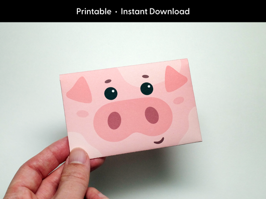 Hand holding a cute, pink pig-themed printable gift card holder designed as an instant digital download. The gift card envelope features a cartoon pig face with big black eyes, floppy ears, and a smiling snout. This DIY craft is an easy-to-make project, perfect for animal lovers and kids’ party favors.