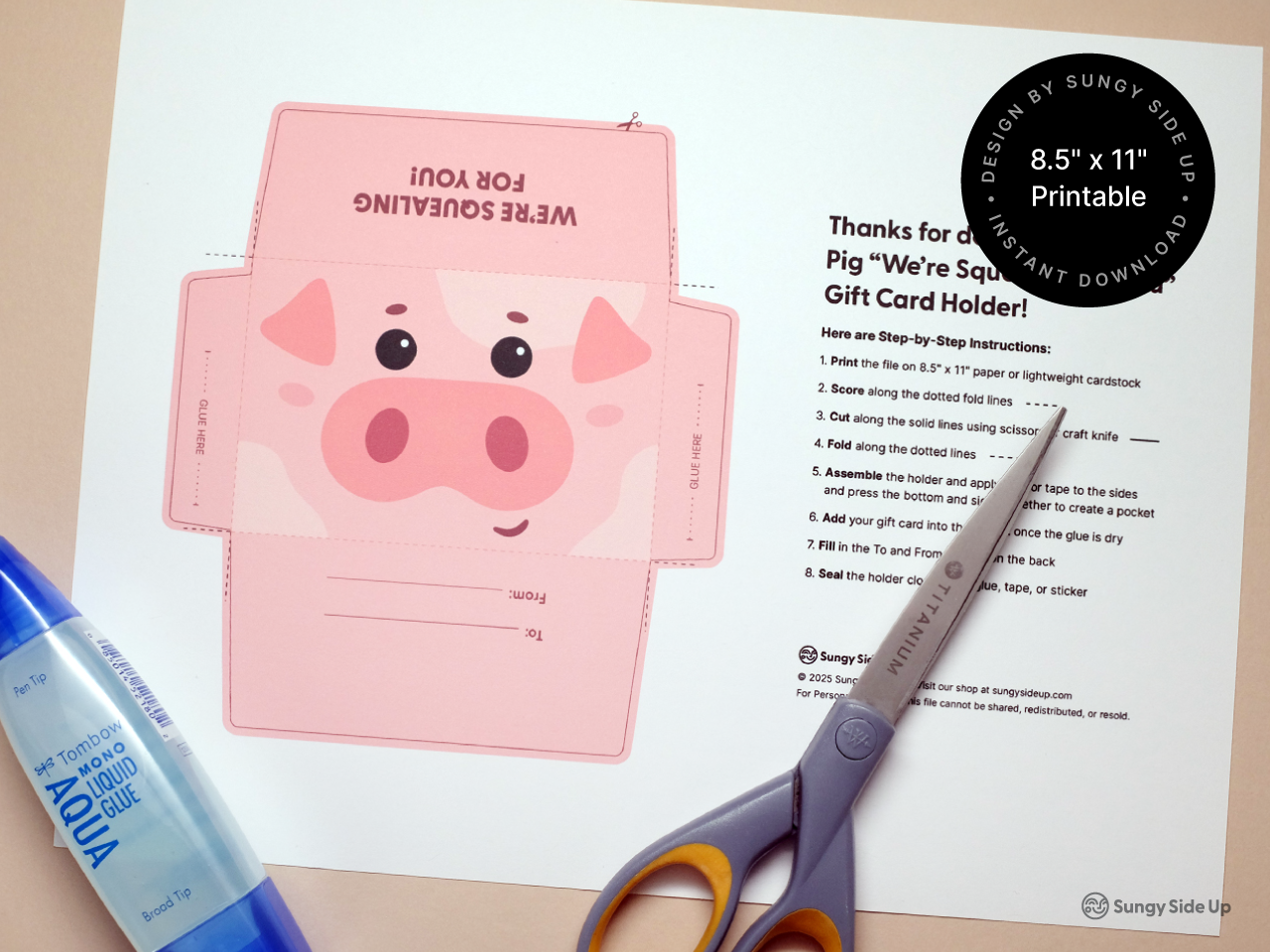 Printable pink pig gift card holder template shown on a US letter sheet of paper, featuring a cartoon pig face with large snout, eyes, and floppy ears. The paper includes cut, fold, and glue lines for easy assembly. A pair of silver craft scissors points to the printed step-by-step instructions on the sheet, emphasizing the DIY aspect of this instant download. Great for parties, birthdays, or creative gift-giving projects.