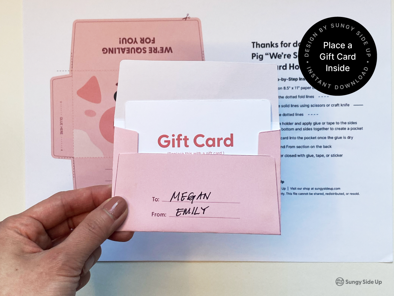 A hand holding a pink printable pig gift card holder with a white gift card inserted. The gift card holder features a To and From section with handwritten names, and the background shows the printed template with the cartoon pig design and assembly instructions. This DIY pig gift card holder is an instant download for birthdays, kids' parties, or animal-themed gifts, combining fun design with easy crafting.
