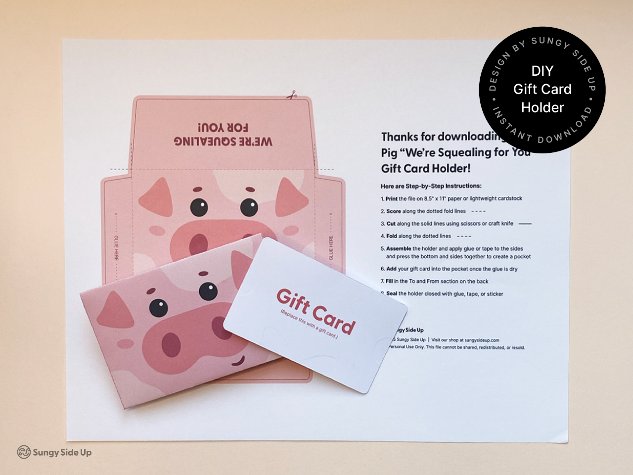 A printable pink pig gift card holder design displayed on a flat surface, next to a white gift card. The gift card holder features a cartoon pig face with floppy ears and a large pink snout, and is partially assembled with clear fold lines. The printable template is part of an instant download DIY craft kit, perfect for birthdays, kids’ parties, or animal-themed events. Includes step-by-step instructions for assembly.