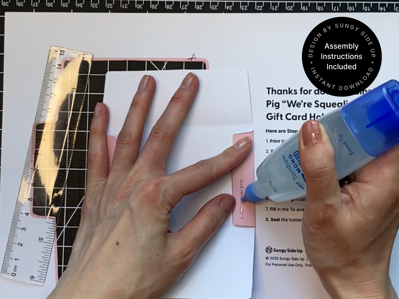 Hands using a glue pen to assemble a printable pig gift card holder, following step-by-step instructions on the background paper. A ruler is used to align the folds precisely. The pink pig gift card template features a cartoon pig face and is being glued along a tab marked "GLUE HERE." This DIY printable is an instant download for easy crafting at home, perfect for animal-themed gifts and party favors.