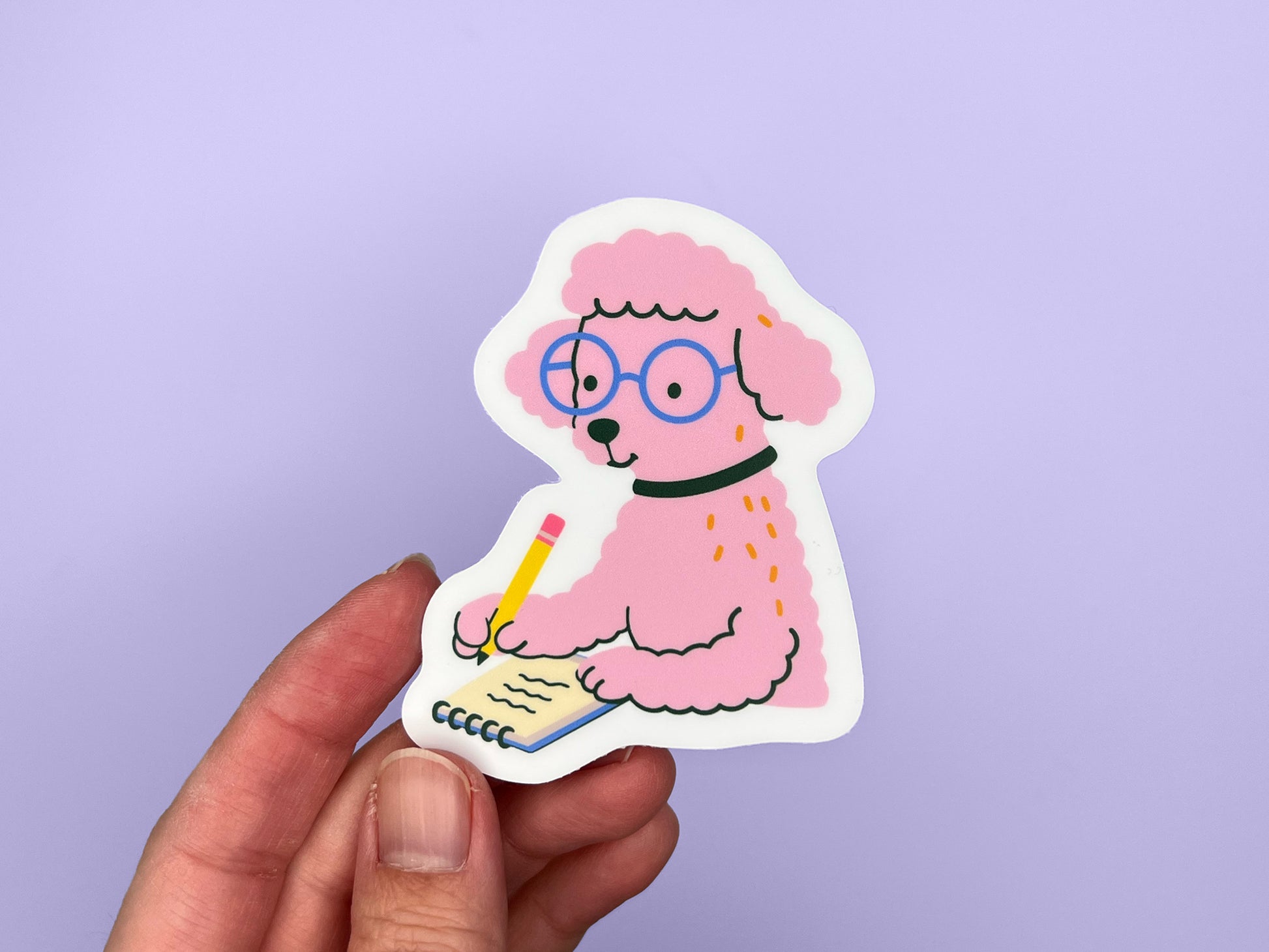 A hand holds a matte die-cut vinyl sticker of a pink illustrated poodle wearing round blue glasses and a black collar while writing in a small spiral notebook with a yellow pencil. The poodle has fluffy textured fur and a calm, focused expression. The sticker has a white border and is shown against a soft purple background.