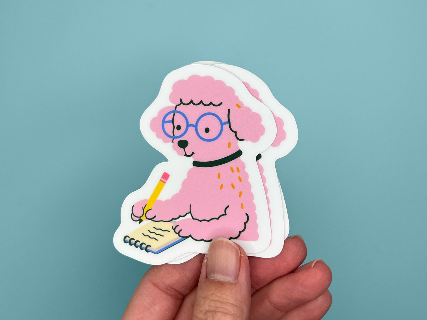 A hand holds three matte die-cut vinyl stickers stacked slightly offset, each showing a pink illustrated poodle wearing round blue glasses and a black collar while writing in a small spiral notebook with a yellow pencil. The poodle has fluffy textured fur and a calm, focused expression. The stickers have white borders and are shown against a teal blue background.