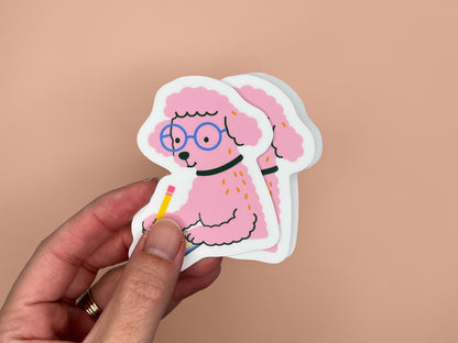 A hand holds two matte die-cut vinyl stickers slightly stacked, each showing a pink illustrated poodle wearing round blue glasses and a black collar while writing in a small spiral notebook with a yellow pencil. The poodle has fluffy textured fur and a calm, focused expression. The stickers have white borders and are shown against a warm peach background.