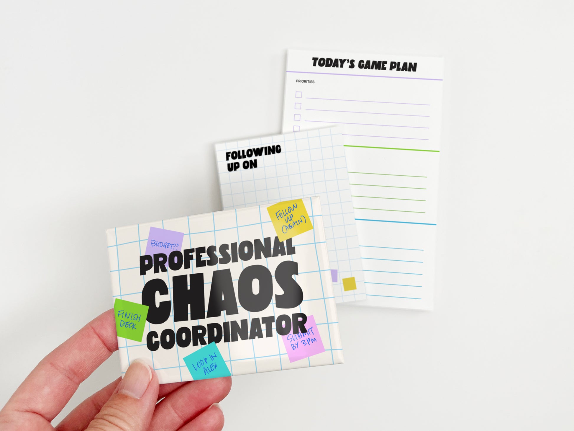 Hand holding a Professional Chaos Coordinator magnet with matching follow-up notepad and Today's Game Plan task notepad in the background.