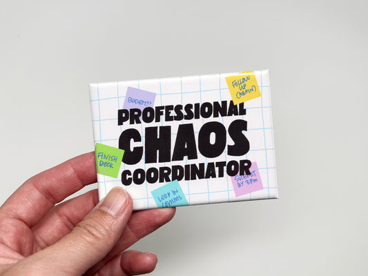 Hand holding a rectangular “Professional Chaos Coordinator” magnet with bold black text, a white grid background, and colorful sticky notes featuring handwritten office reminders.