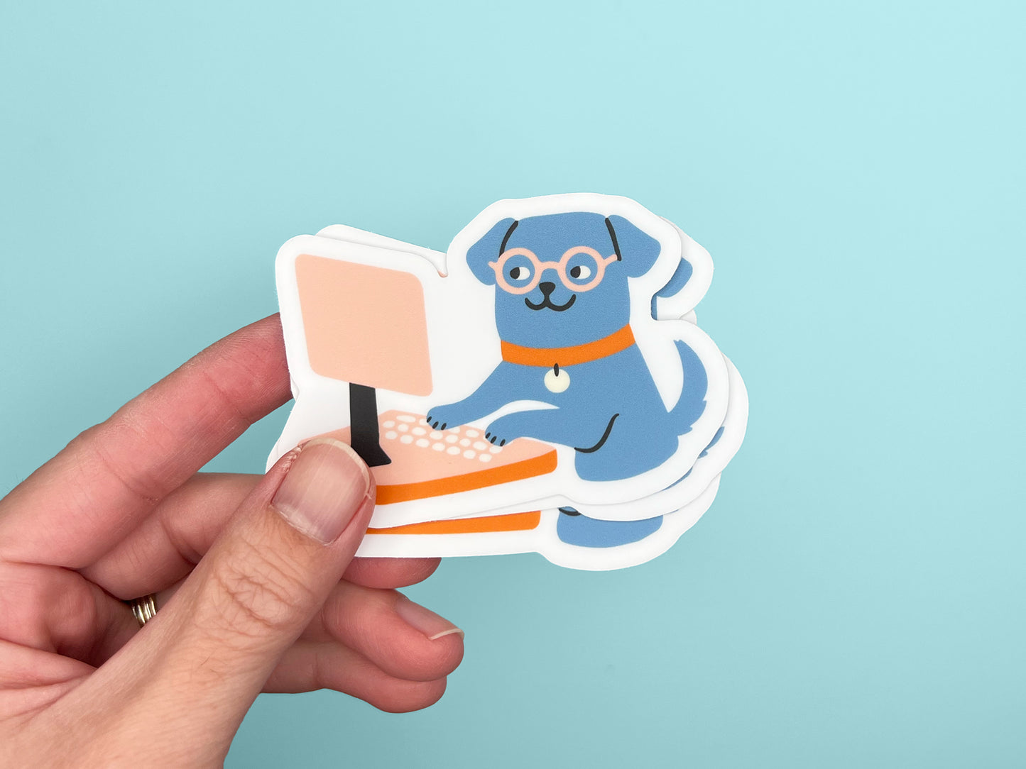 Hand holding a small stack of matte die cut vinyl stickers featuring an illustrated blue dog wearing round glasses and an orange collar, typing on a peach-colored computer. Stickers have white borders and are shown against a light teal background.
