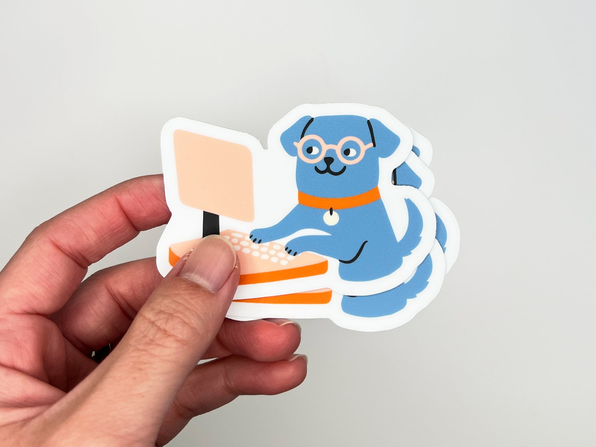 Hand holding a small stack of matte die cut vinyl stickers showing an illustrated blue dog wearing round glasses and an orange collar, typing on a peach-colored desktop computer. Stickers have white borders and are photographed against a light neutral background.