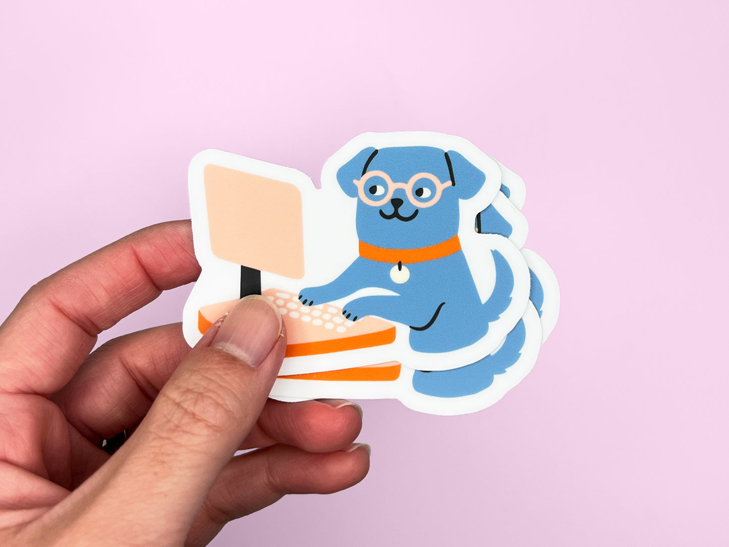 Hand holding a small stack of matte die cut vinyl stickers showing an illustrated blue dog wearing round glasses and an orange collar, typing on a peach-colored desktop computer. Stickers have white borders and are photographed against a soft pink background.
