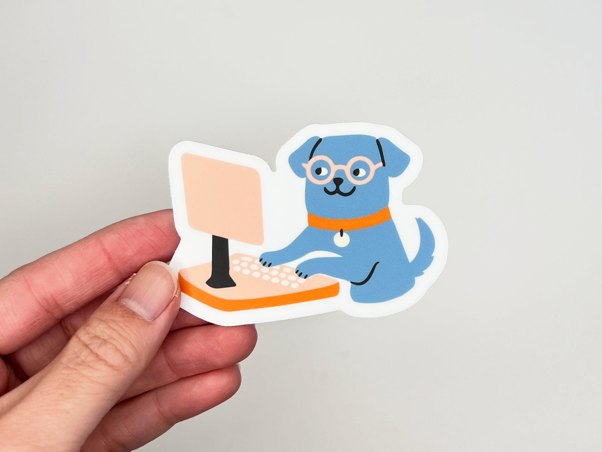 Hand holding a single matte die cut vinyl sticker of an illustrated blue dog wearing round glasses and an orange collar, sitting upright and typing on a peach-colored desktop computer. Sticker has a white border and is photographed against a light neutral background.