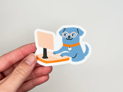 Hand holding a single matte die cut vinyl sticker of an illustrated blue dog wearing round glasses and an orange collar, sitting upright and typing on a peach-colored desktop computer. Sticker has a white border and is photographed against a light neutral background.