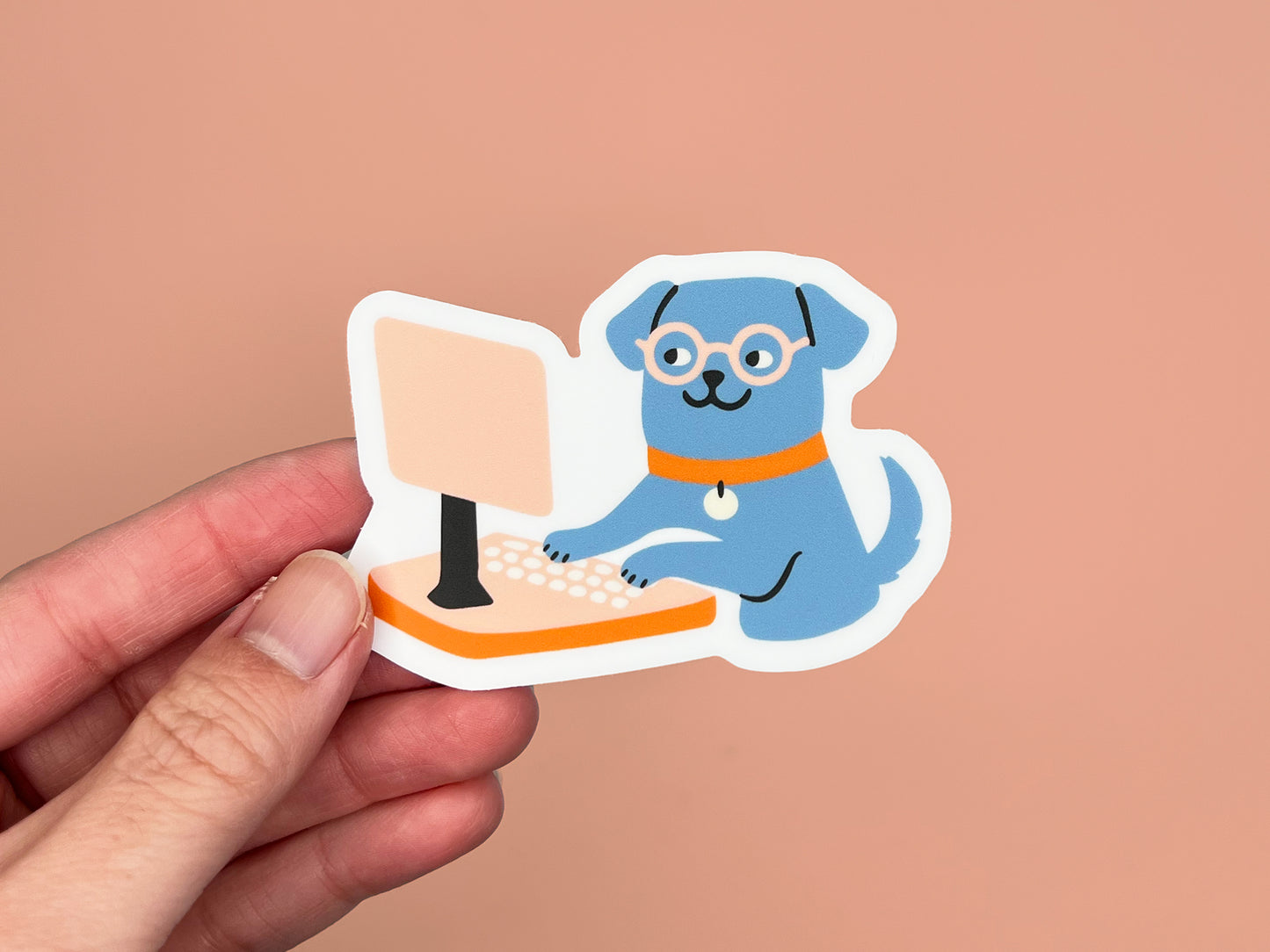 Hand holding a matte die cut vinyl sticker of an illustrated blue dog wearing round glasses and an orange collar, typing on a peach-colored desktop computer. Sticker has a white border and is photographed against a warm peach background.