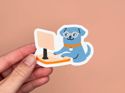 Hand holding a matte die cut vinyl sticker of an illustrated blue dog wearing round glasses and an orange collar, typing on a peach-colored desktop computer. Sticker has a white border and is photographed against a warm peach background.