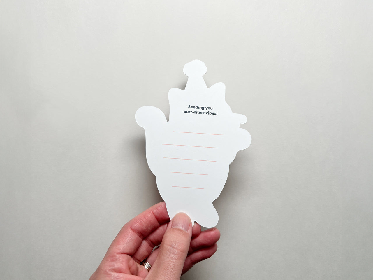 A hand holds the back of a die-cut note card shaped like a cat. The back has lined spaces for writing and the printed message Sending you purr-sitive vibes! at the top. The background is a neutral light gray color.