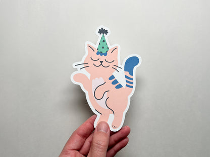 A hand holds the front of a die-cut note card shaped like a pink cat wearing a green and black party hat. The cat has a happy, playful expression and a blue tail. The background is a neutral light gray color.