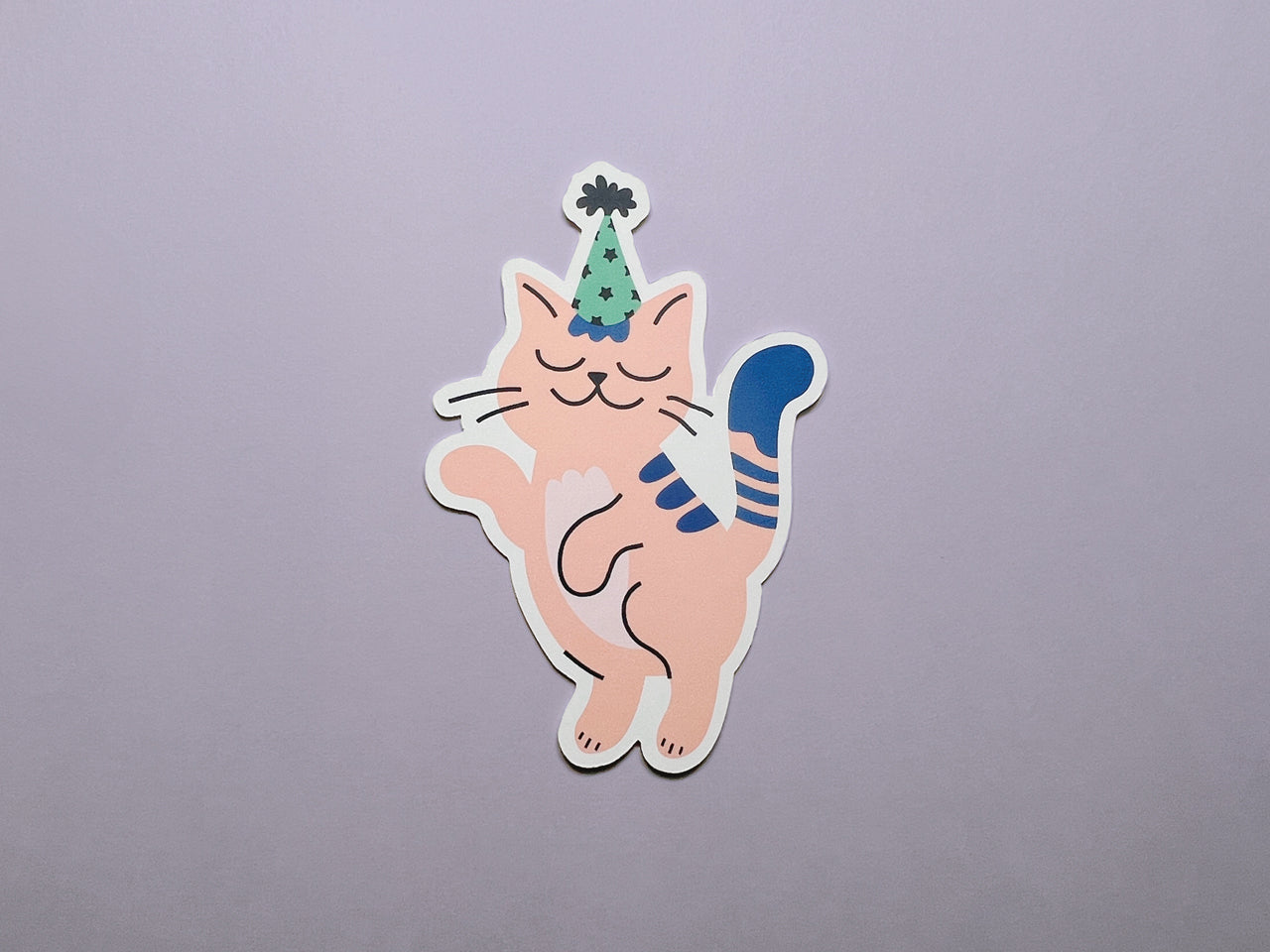 A die-cut note card featuring a pink cat wearing a green and black party hat is placed on a flat surface with a soft purple background. The design of the card shows an illustration of a cat in a joyful, dancing pose with a blue tail and paws.