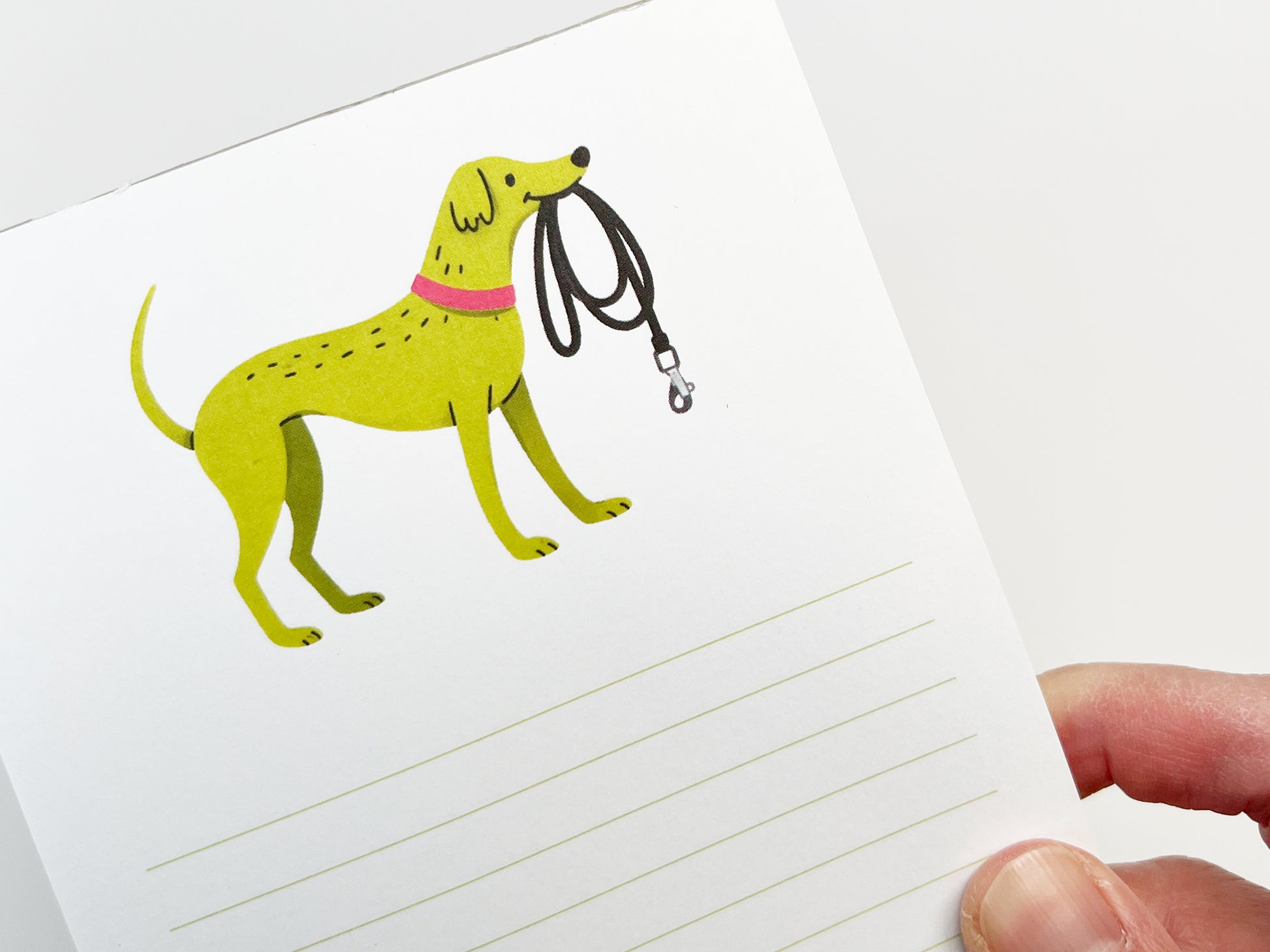 A close-up of a 4.25 x 5.5 inch lined notepad featuring an illustrated green dog holding a black leash in its mouth, wearing a pink collar. The notepad is held at an angle by a hand against a clean white background, with light green writing lines visible below the illustration.