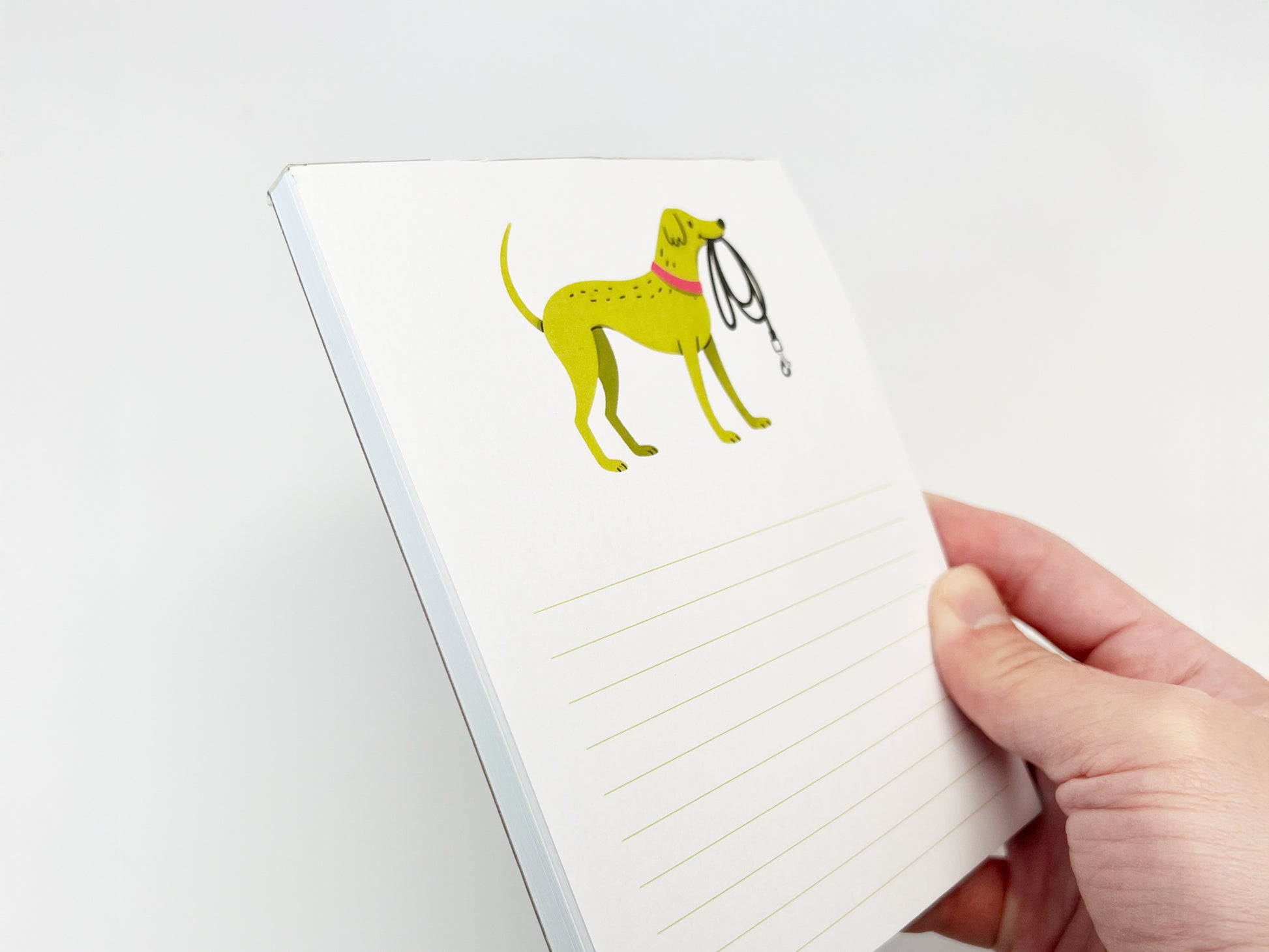 A hand holding a small 4.25 x 5.5 inch lined notepad at an angle, showing the thickness of the paper stack. The notepad features an illustrated green dog wearing a pink collar and holding a black leash in its mouth near the top, with light green writing lines below on a white page and a clean white background.