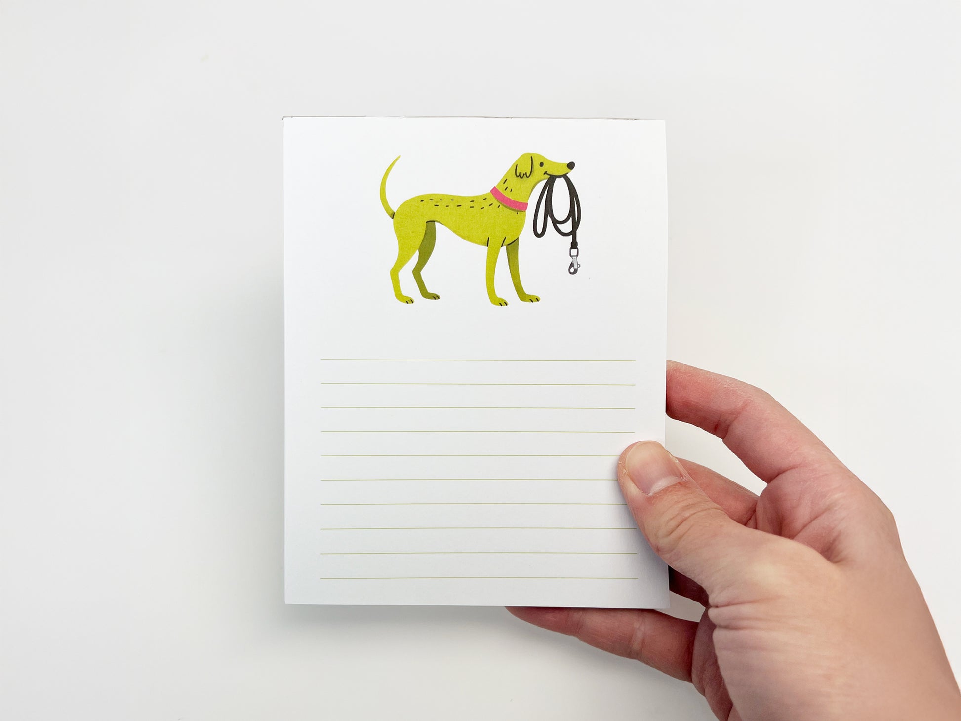 A hand holding a small 4.25 x 5.5 inch lined notepad upright against a white background. The notepad features an illustrated green dog wearing a pink collar and holding a black leash in its mouth at the top, with light green horizontal writing lines filling the lower portion of the page.