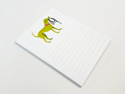 A small 4.25 x 5.5 inch lined notepad lying flat on a white surface. The top of the page features an illustrated green dog wearing a pink collar and holding a black leash, with light green horizontal writing lines on the right side of the page and a visible stack of paper sheets beneath.
